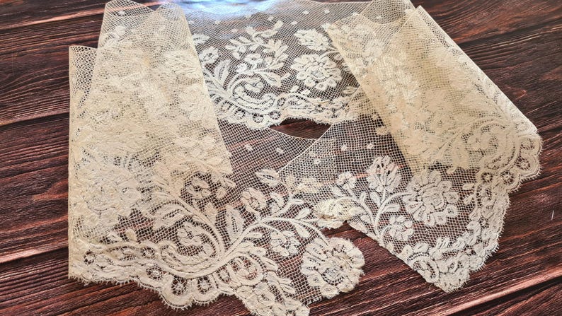 Puede incluir: Tela de encaje color marfil con dise&ntilde;os florales y de hojas. El encaje se muestra sobre una superficie de madera oscura, resaltando su delicado dise&ntilde;o y bordes festoneados. La tela est&aacute; doblada en secciones.
