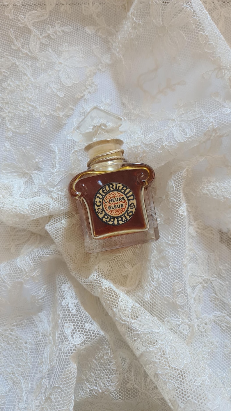 May include: A vintage glass bottle of Guerlain L'Heure Bleue perfume. The bottle is clear glass with a gold-colored cap and a label with the text "Guerlain L'Heure Bleue Paris".