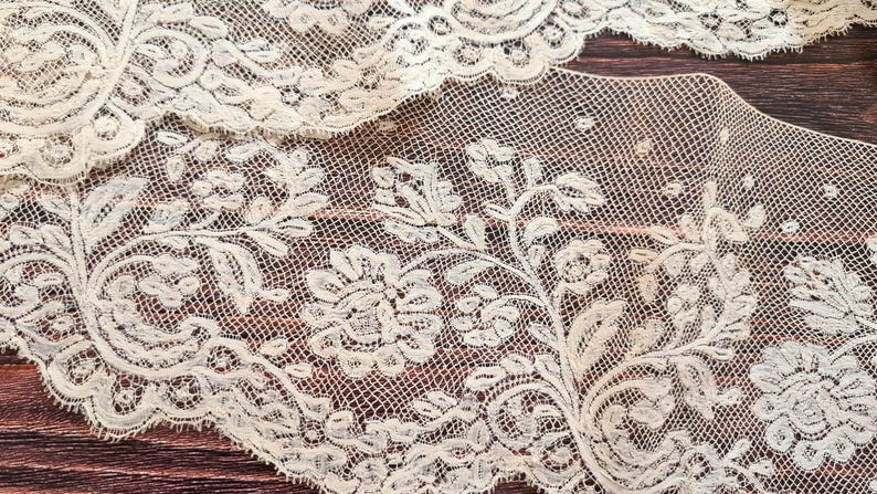 Puede incluir: Primer plano de tela de encaje color crema con dise&ntilde;os florales y de hojas. El encaje tiene un tejido delicado y abierto y est&aacute; colocado sobre una superficie de madera oscura. El dise&ntilde;o presenta motivos detallados y repetidos.