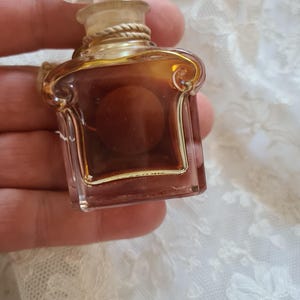 May include: A small, clear glass bottle of perfume with a gold cap and a gold rope detail around the neck. The bottle is filled with a brown liquid.