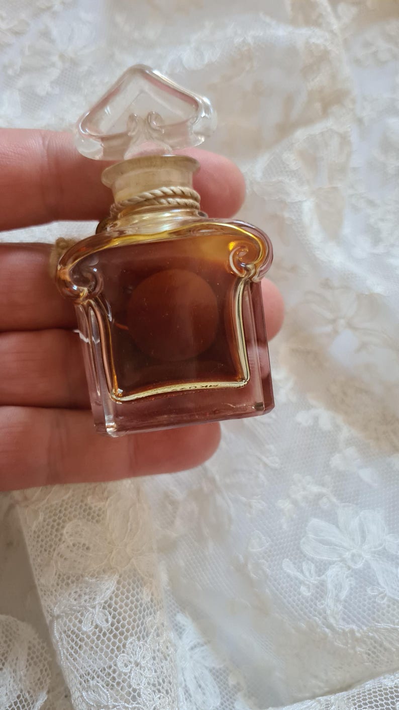 May include: A small, clear glass bottle of perfume with a gold cap and a gold rope detail around the neck. The bottle is filled with a brown liquid.