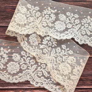 Puede incluir: Dos piezas de encaje marfil con motivos florales y de hojas. El encaje tiene un borde festoneado y est&aacute; sobre un fondo de vetas de madera oscura. El encaje probablemente se utiliza para coser o hacer manualidades.