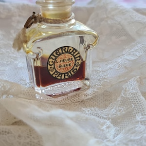 May include: A vintage glass bottle of perfume with a gold cap and a label that reads "Guerlain L'Heure Bleue Paris". The bottle is filled with a dark brown liquid and is sitting on a white lace fabric.