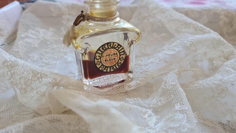 May include: A vintage glass bottle of perfume with a gold cap and a label that reads "Guerlain L'Heure Bleue Paris". The bottle is filled with a dark brown liquid and is sitting on a white lace fabric.