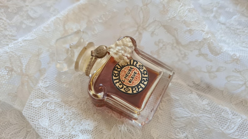 May include: A vintage glass bottle of perfume with a clear glass stopper and a label that reads "Guerlain L'Heure Bleue Paris".