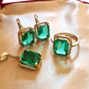 Può includere: Un set di gioielli composto da un ciondolo, un anello e un paio di orecchini. Ogni pezzo ha una montatura in oro e una grande pietra preziosa verde smeraldo a taglio quadrato. Le pietre preziose sono circondate da pietre più piccole e trasparenti.