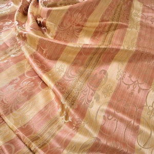 May include: A close-up of a folded fabric with a striped pattern. The fabric has alternating stripes of peach and gold. Floral designs are visible within the stripes, adding texture and detail to the material.