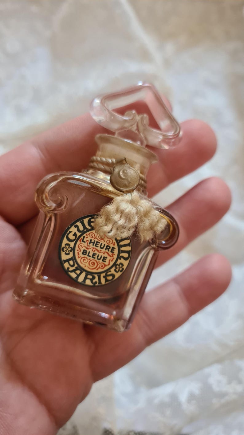 May include: A small, clear glass bottle of perfume with a gold-colored cap and a label that reads "Guerlain L'Heure Bleue Paris".