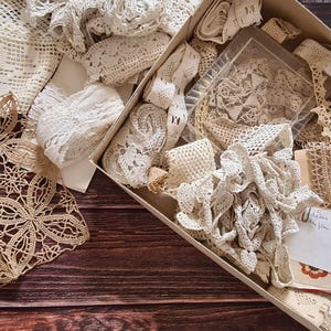 May include: A collection of vintage lace and trim in various shades of cream and beige. The assortment includes doilies, ribbons, and lace pieces, some in a cardboard box. The items are arranged on a dark wooden surface.