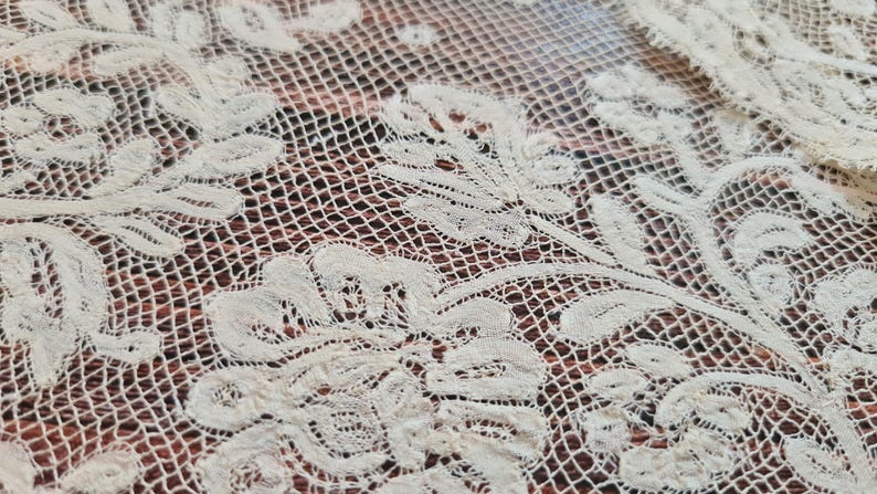 Puede incluir: Primer plano de tela de encaje blanquecina con motivos florales. El encaje presenta dise&ntilde;os florales detallados y en relieve y un delicado fondo de red. La tela probablemente se utiliza con fines decorativos.