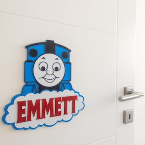 Puede incluir: Un personaje azul y negro de Thomas la locomotora con una cara blanca y letras rojas que dicen "EMMETT" en una nube blanca. El personaje está unido a una puerta blanca.