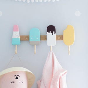 May include: A wooden wall-mounted coat rack featuring four colorful popsicle-shaped hooks. The hooks are painted in pastel colors: pink, blue, and yellow, with a brown wooden base. A pink hat and a pink garment hang from the hooks.