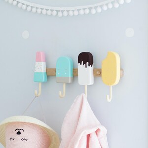 May include: Four colorful popsicle-shaped wall hooks mounted on a wooden board. The hooks are pastel colors: pink, blue, white, and yellow. A pink hat and a pink towel hang from the hooks. White pom-pom garland decorates the wall.