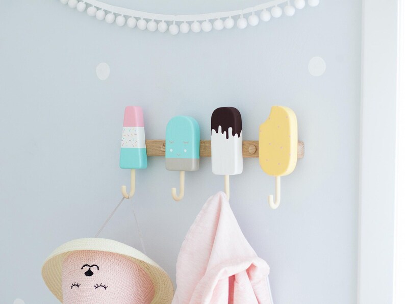 May include: Four colorful popsicle-shaped wall hooks mounted on a wooden board. The hooks are pastel colors: pink, blue, white, and yellow. A pink hat and a pink towel hang from the hooks. White pom-pom garland decorates the wall.