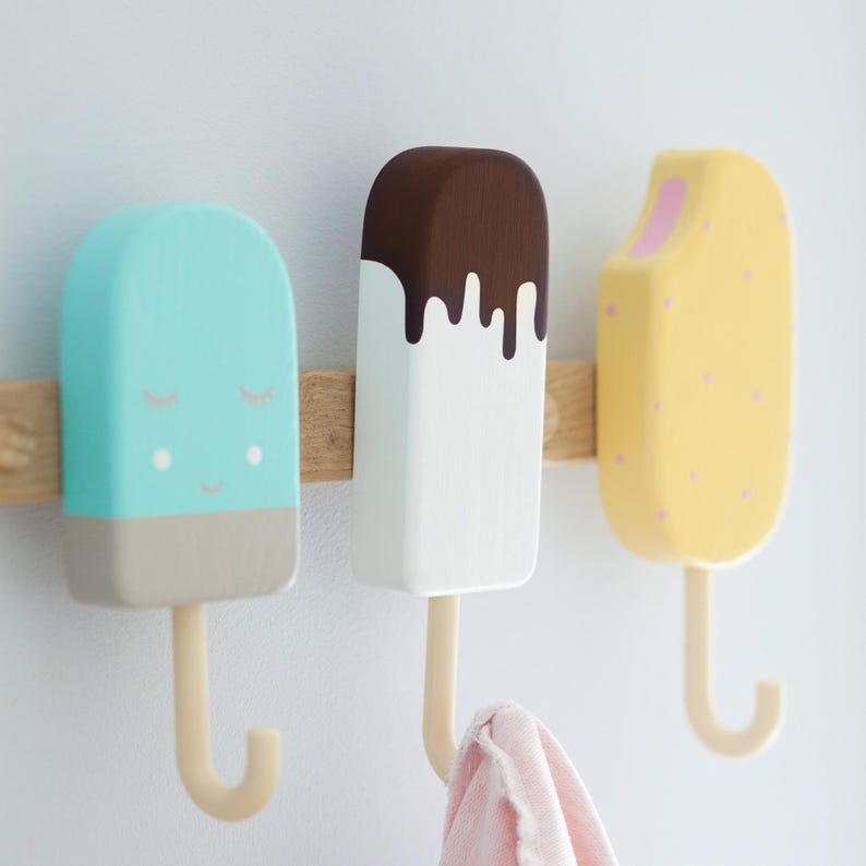 May include: Three wooden ice cream-shaped wall hooks in pastel colors. The hooks are mounted on a wooden bar. One is light blue with a smiling face, one is white with chocolate dripping, and the last is yellow with pink dots.