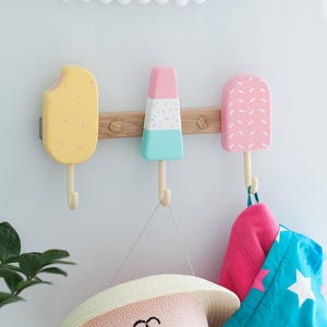 Könnte beinhalten: Drei Holz-Wandhaken in Eis am Stiel-Form, gelb, rosa und blau, an einer Holzleiste befestigt. Ein Hut, ein rosa Handtuch und eine blaue Tasche mit weißen Sternen hängen daran. Eine verspielte Wanddekoration.