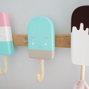 May include: Three colorful popsicle-shaped wall hooks mounted on a wooden board. The hooks are painted in pastel colors: pink, blue, and white, with decorative details. Each hook has a wooden stick and a curved hook for hanging items.