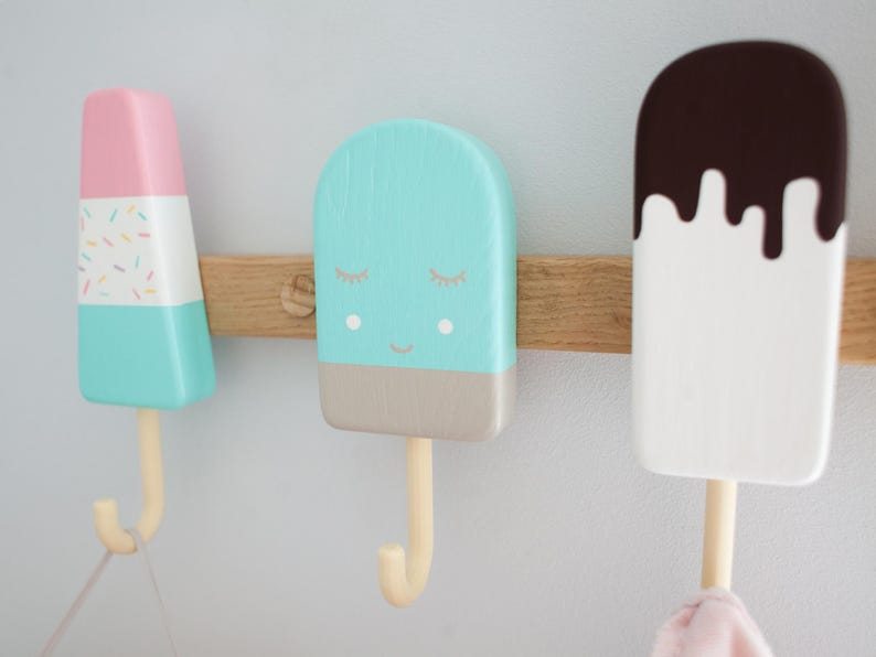 May include: Three colorful popsicle-shaped wall hooks mounted on a wooden board. The hooks are painted in pastel colors: pink, blue, and white, with decorative details. Each hook has a wooden stick and a curved hook for hanging items.