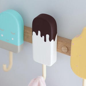 May include: Three wooden popsicle-shaped wall hooks in pastel colors. One is light blue with a gray base and a smiling face, another is white with a brown top, and the last is yellow with pink dots. They are mounted on a wooden plank.