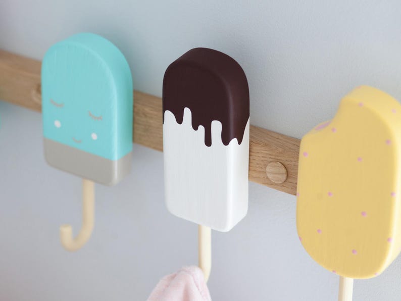 May include: Three wooden popsicle-shaped wall hooks in pastel colors. One is light blue with a gray base and a smiling face, another is white with a brown top, and the last is yellow with pink dots. They are mounted on a wooden plank.