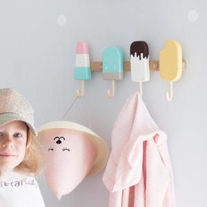 May include: A wall-mounted coat rack featuring four colorful popsicle-shaped hooks. The hooks are in pastel colors: pink, blue, white, and yellow. A pink hat with a face design and a pink towel hang from the hooks.