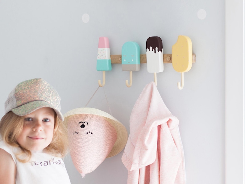 May include: A wall-mounted coat rack featuring four colorful popsicle-shaped hooks. The hooks are in pastel colors: pink, blue, white, and yellow. A pink hat with a face design and a pink towel hang from the hooks.