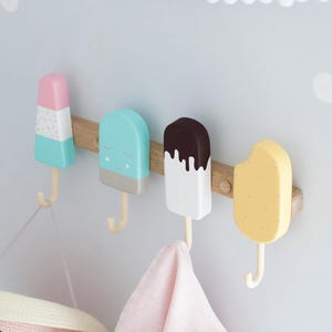 May include: A wooden wall-mounted coat rack featuring four colorful popsicle-shaped hooks. The hooks are in pastel colors: pink, blue, white, and yellow, with brown wooden accents. A pink towel and a hat hang from the hooks.