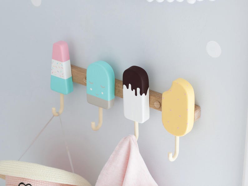 May include: A wooden wall-mounted coat rack featuring four colorful popsicle-shaped hooks. The hooks are in pastel colors: pink, blue, white, and yellow, with brown wooden accents. A pink towel and a hat hang from the hooks.