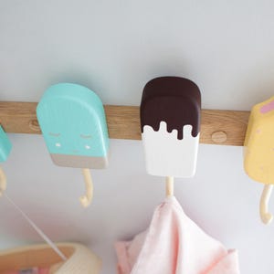 May include: A wooden wall-mounted coat rack featuring four popsicle-shaped hooks in pastel colors. The hooks are painted pink, blue, white, and yellow, with details like sprinkles and melting chocolate. A pink cloth hangs from one hook.