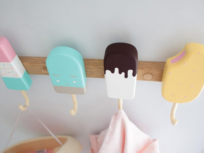 May include: A wooden wall-mounted coat rack featuring four popsicle-shaped hooks in pastel colors. The hooks are painted pink, blue, white, and yellow, with details like sprinkles and melting chocolate. A pink cloth hangs from one hook.
