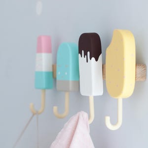 May include: Four colorful popsicle-shaped wall hooks. The hooks are painted in pastel colors: pink, blue, white, and yellow. Each hook has a wooden base and a cream-colored hook. A pink towel hangs from one hook.