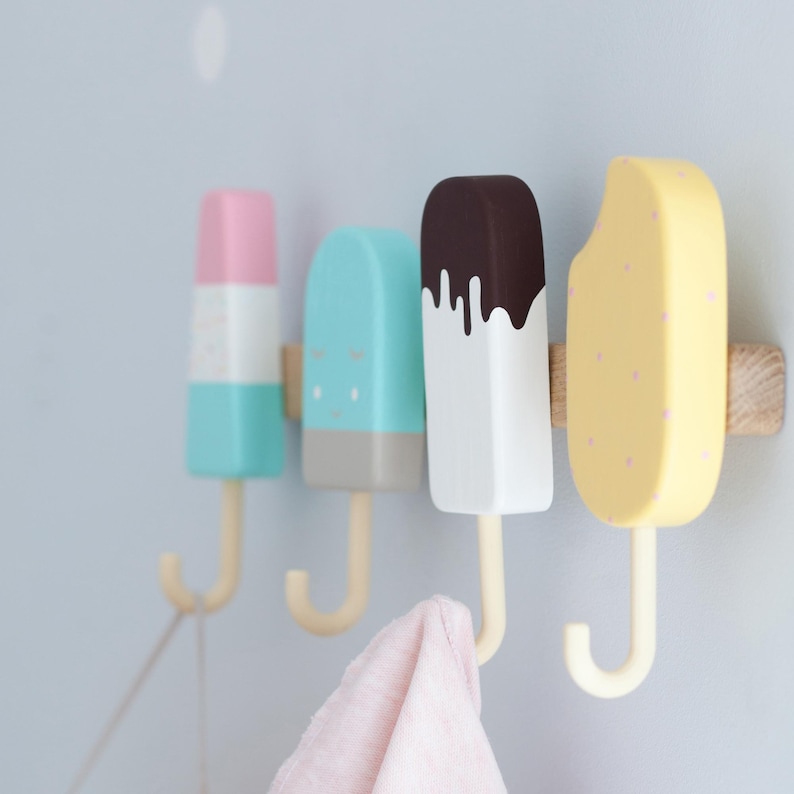 May include: Four colorful popsicle-shaped wall hooks. The hooks are painted in pastel colors: pink, blue, white, and yellow. Each hook has a wooden base and a cream-colored hook. A pink towel hangs from one hook.