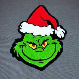 Op de afbeelding: Groen Grinch-gezicht met een rood-witte kerstmanmuts. De Grinch glimlacht met een ondeugende blik in zijn ogen.