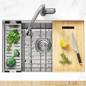 Puede incluir: Un fregadero de cocina de acero inoxidable con una tabla de cortar integrada y un grifo extra&iacute;ble. El fregadero tiene un desag&uuml;e de rejilla y est&aacute; lleno de agua. Hay verduras y un lim&oacute;n en la tabla de cortar.