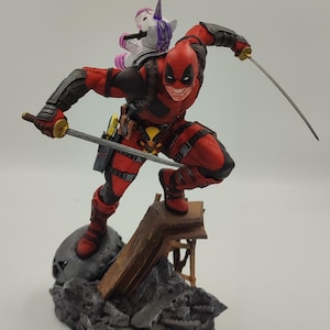 May include: An action figure of Deadpool, dressed in red and black, holding two swords. A small pink unicorn figure is on his back. The figure is standing on a grey and brown rock base.