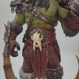 Puede incluir: Una figura de guerrero Orc verde con un hacha grande, vistiendo un atuendo de cuero y piel marrón con una hebilla de cinturón de calavera. La figura está de pie sobre una base rocosa.