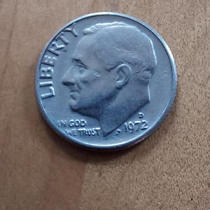 Könnte beinhalten: Eine silberne US-amerikanische 10-Cent-Münze von 1972 mit einem Profil von Franklin D. Roosevelt und den Inschriften "LIBERTY" und "IN GOD WE TRUST". Die Münze liegt auf einer Holzoberfläche.