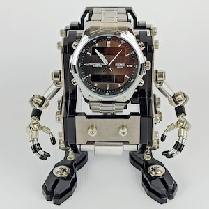 Può includere: Un orologio SKMEI argento e nero è montato su un supporto a forma di robot. L'orologio ha un quadrante scuro con lancette e indicatori argentati. Il supporto robot ha braccia e gambe articolate, con componenti neri e argentati.