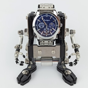 May include: A silver wristwatch with a blue face is mounted on a black and silver robot sculpture. The robot has articulated arms and legs, with a mechanical aesthetic. The watch features multiple subdials and a stainless steel strap.