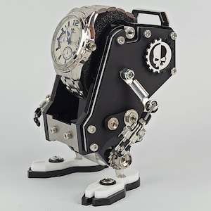 May include: A silver wristwatch with a white face and blue hands is displayed on a black and silver mechanical stand. The stand has a skull logo and a gear design. The base and feet are white.
