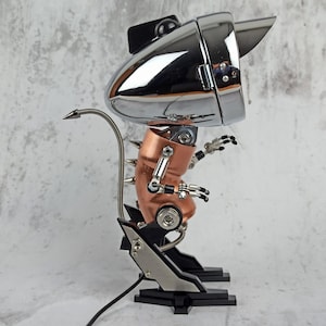 Może przedstawiać: Robotyczna rzeźba wykonana z chromu i miedzi z długim, spiczastym ogonem. Robot ma dużą, okrągłą głowę z chromowanym reflektorom i miedziane ciało z ruchomymi kończynami.