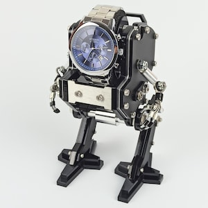 Peut inclure: Une montre-bracelet argentée avec un cadran bleu est montée sur un support noir et argenté en forme de robot. Le support a des bras et des jambes articulés, lui donnant un aspect mécanique. La montre est le point central de cet affichage unique.