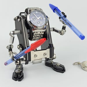 Puede incluir: Un reloj de pulsera plateado con esfera azul es sostenido por un dispositivo mecánico negro y plateado. Dos bolígrafos, uno rojo y otro azul, son sostenidos por el dispositivo. El dispositivo tiene brazos articulados y una base con abrazaderas ajustables.