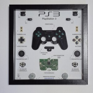Könnte beinhalten: Gerahmtes Kunstwerk mit PlayStation 3 Controller-Komponenten. Der schwarze Rahmen umgibt einen weißen Hintergrund mit einem schwarzen Controller, einer Hauptplatine und anderen Teilen. Text enthält "PS3 PlayStation 3", "SONY" und Komponentenbezeichnungen.