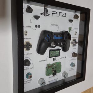 Könnte beinhalten: Gerahmte Präsentation eines zerlegten PS4-Controllers, der seine Komponenten zeigt. Der schwarze Controller ist teilweise zerlegt und zeigt interne Teile wie die Hauptplatine und den Lautsprecher. Beschriftete Komponenten umfassen Tasten, Joysticks und die Frontschale, alles vor weißem Hintergrund.