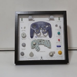 Puede incluir: Exhibición enmarcada de un mando de Nintendo GameCube desmontado. Los componentes incluyen el mando morado, la placa base, los botones y otras piezas. El marco negro tiene un fondo blanco con etiquetas para cada pieza.