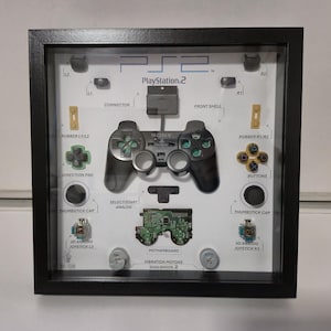May include: A framed PlayStation 2 controller deconstructed, showcasing individual components like buttons, joystick, and motherboard. The black frame surrounds a white background with the controller's parts labeled. The PlayStation 2 logo is visible at the top.