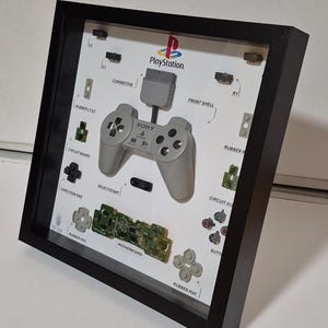 Puede incluir: Una exhibición enmarcada que presenta un mando de PlayStation desmontado. Los componentes, incluido el mando, las placas de circuito y los botones, están dispuestos sobre un fondo blanco. El logotipo de PlayStation es visible. El marco es negro.