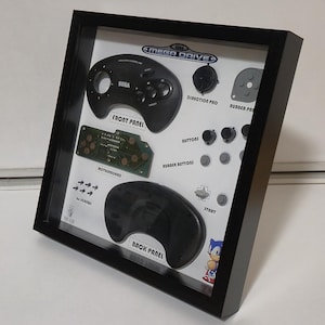 Könnte beinhalten: Gerahmte Anzeige von Sega Mega Drive Controller-Teilen. Der schwarze Rahmen umgibt einen weißen Hintergrund mit Frontplatte, Motherboard und Rückplatte. Enthält Richtungspad, Tasten und das Sega-Logo.