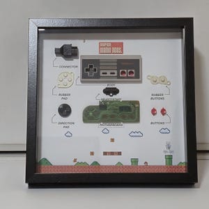 May include: Framed artwork featuring a deconstructed Nintendo controller with labeled parts. The design includes the "Super Mario Bros." logo, a controller body, motherboard, buttons, and a pixelated landscape. The frame is black.